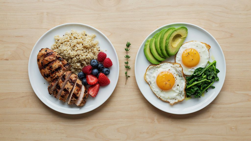 Split overhead photo comparing a moderate low-carb plate with grilled chicken, quinoa, and berries on one side and a strict ketogenic plate with avocado, eggs, and leafy greens on the other