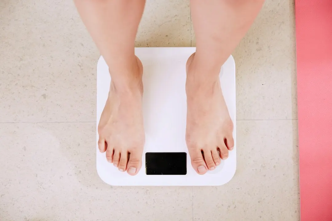 A person looking at a bathroom digital scale neutrally while holding a soft tape measure around their waist.